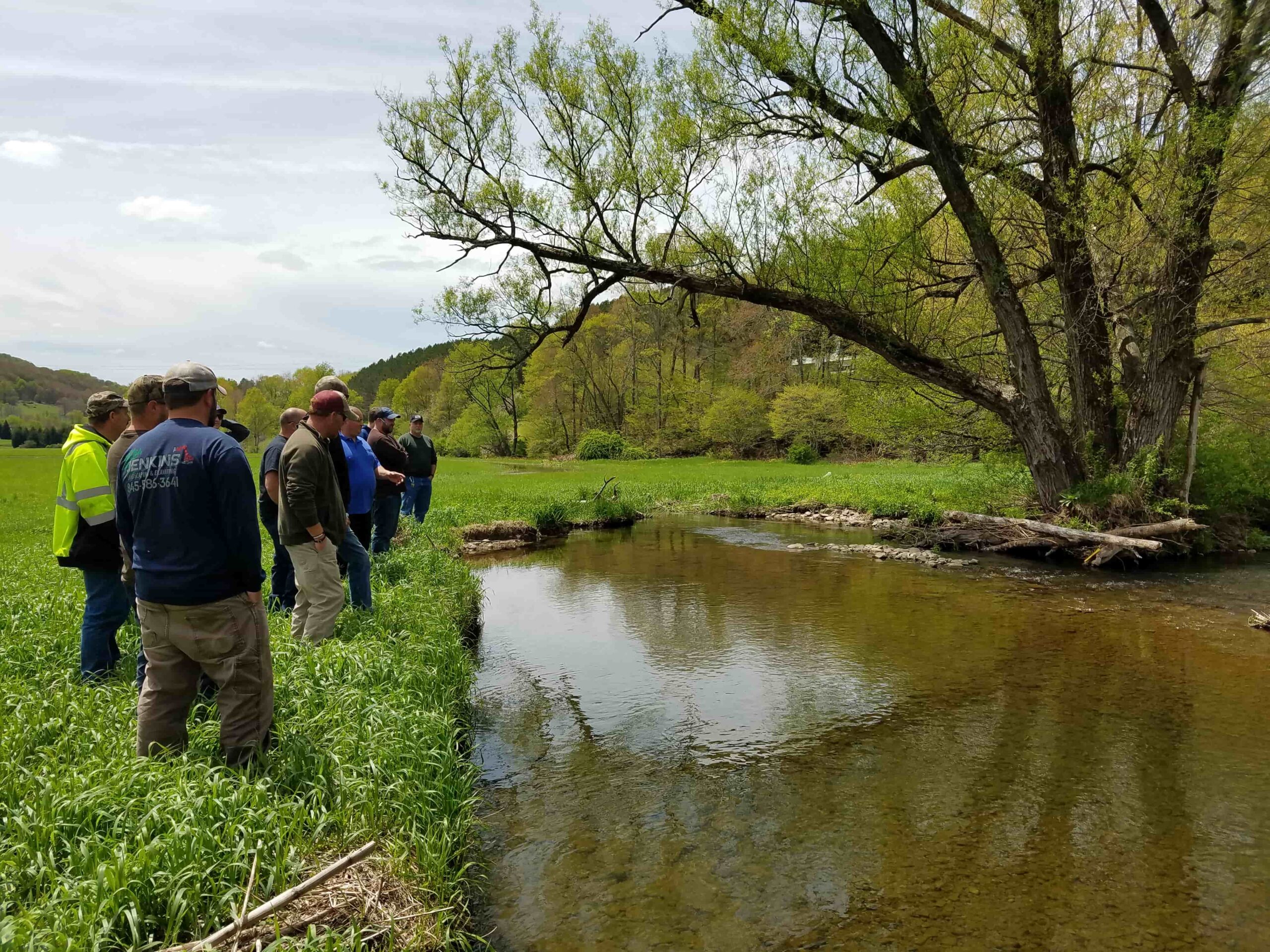 Image shows group of individuals standing in green field overlooking stream.