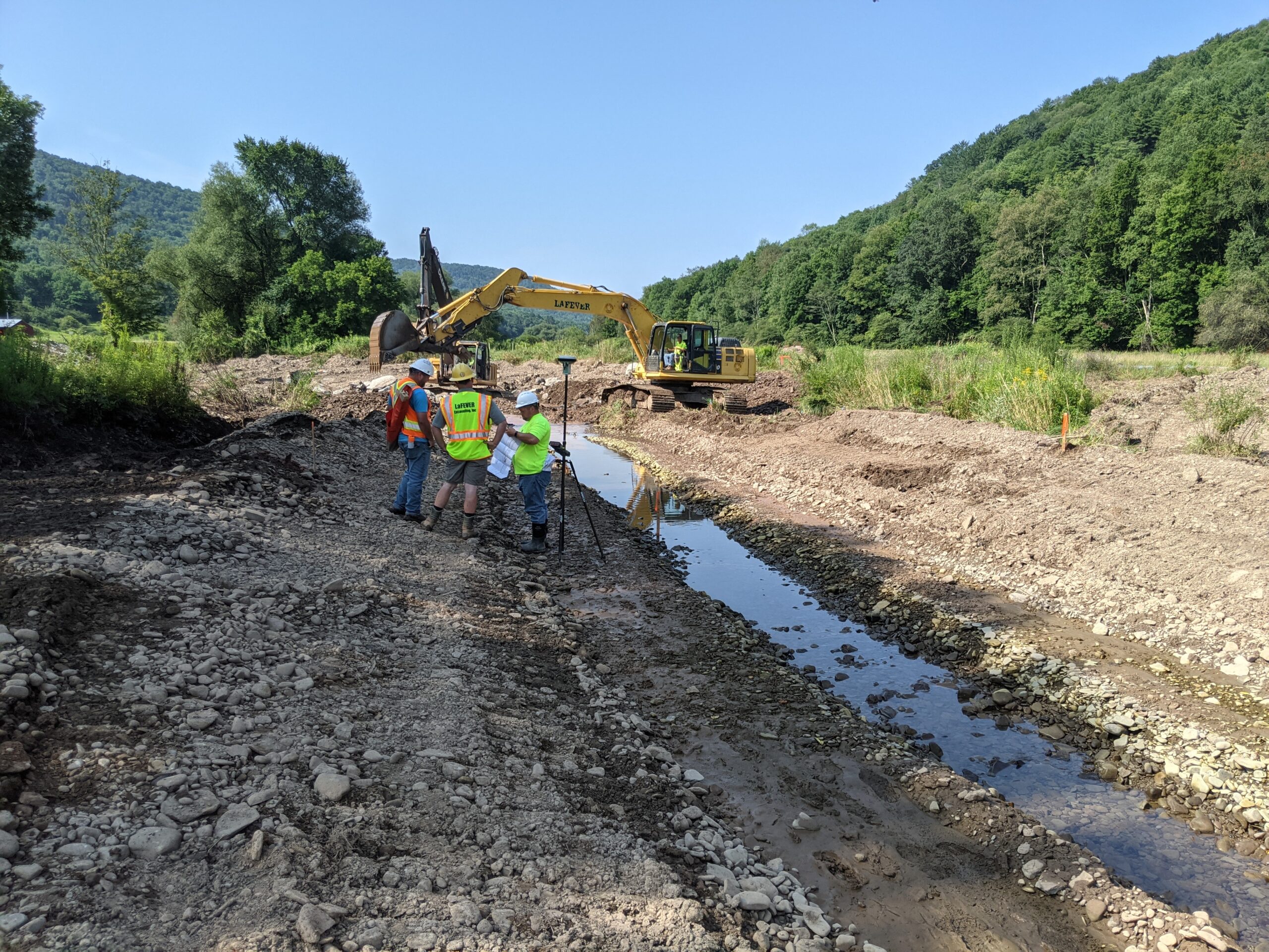 Image of construction and survey of East Brook Stream 20210809-H002 by three individuals standing next to stream reviewing paperwork. Excavator in background with operator inside. Landscape is stream in valley surrounded by active worksite and green forested hills.