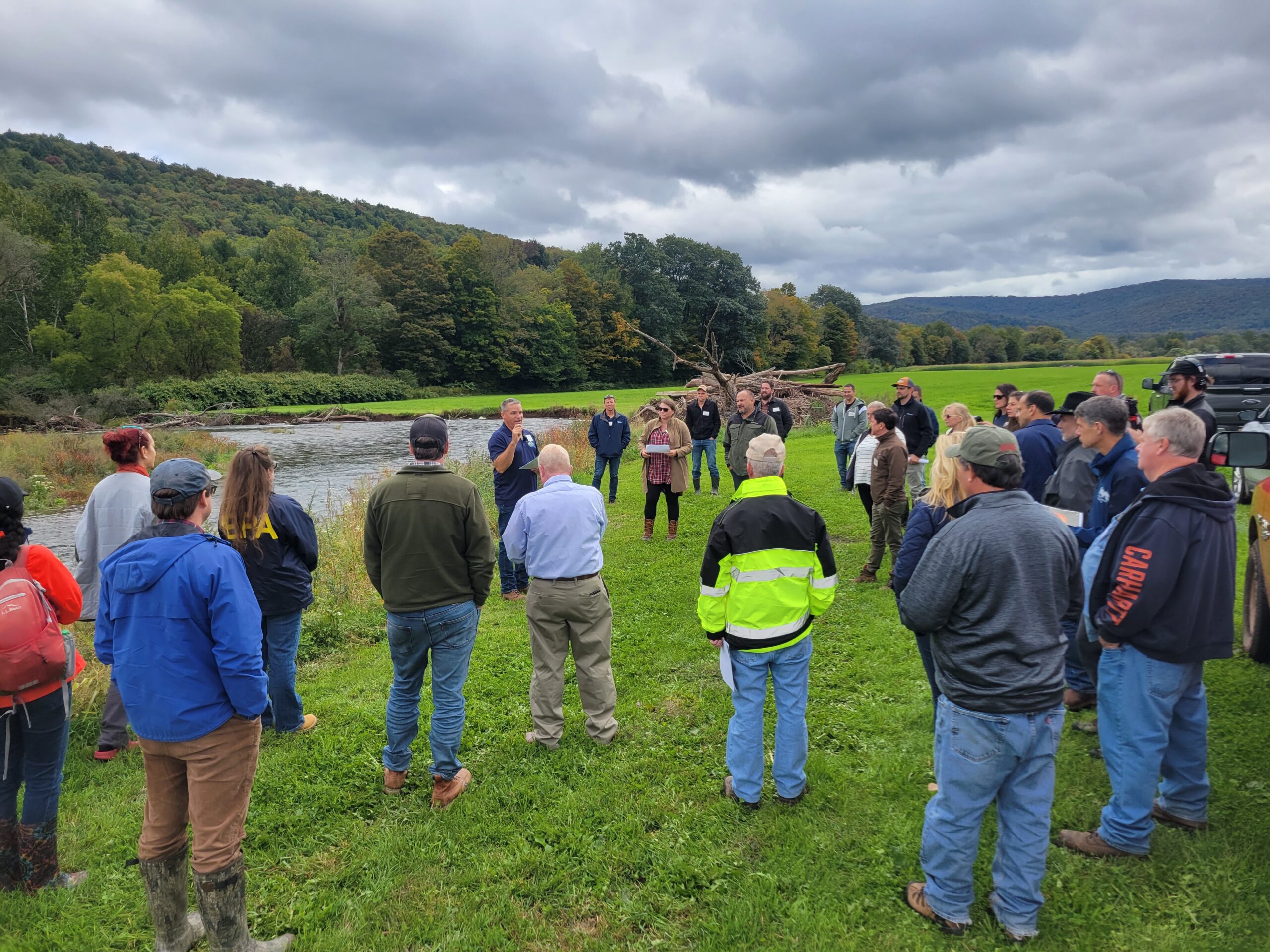 Image description: DCSWCD's Stream Program Coordinator Graydon Dutcher presents to gathering of local and state officials and organizations about West Branch of the Delaware River project. Group is standing in green field adjacent to river with forested mountains in background.